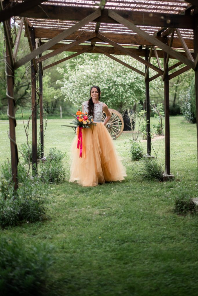 Mariée en robe tulle orange tenant un bouquet coloré sous une pergola en bois dans un jardin des Landes