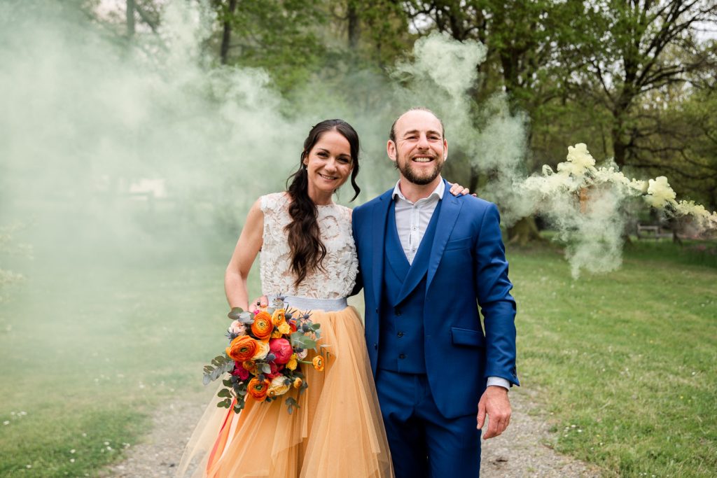 Couple de mariés souriant entouré de fumée colorée lors d’un shooting mariage en extérieur
